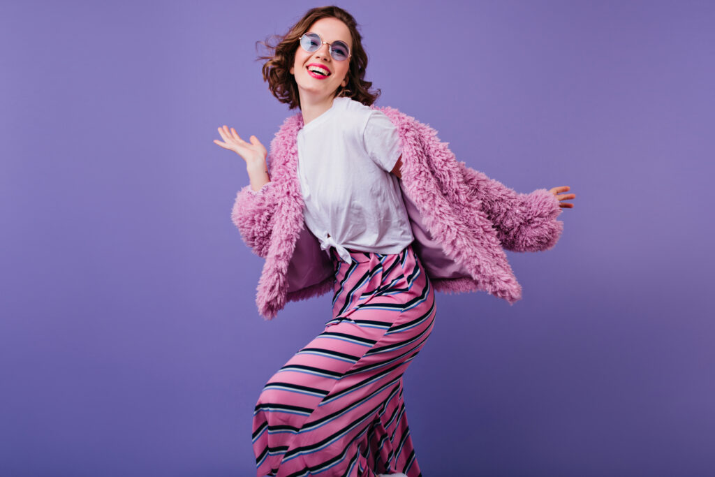 Blissful curly girl in sunglasses laughing while posing in pink fur jacket. Indoor shot of cute young woman dancing on purple background with smile.