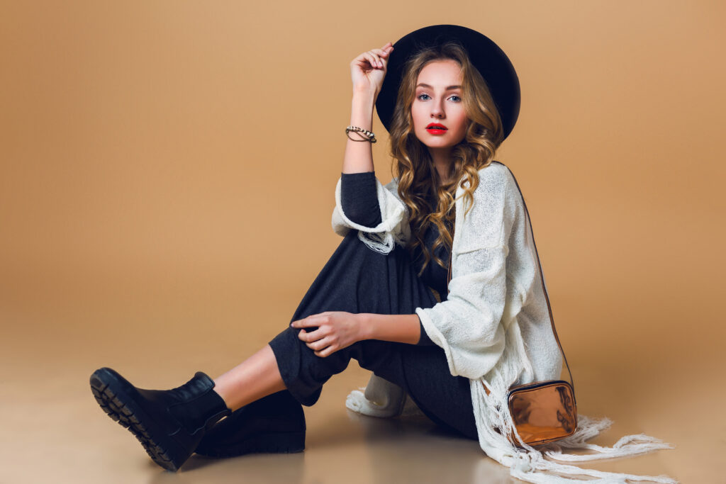 High fashion portrait of young elegant blonde  woman in black wool hat  wearing oversize white fringe  poncho with long grey dress. Studio shot. American hippie bohemian style. Beige background.