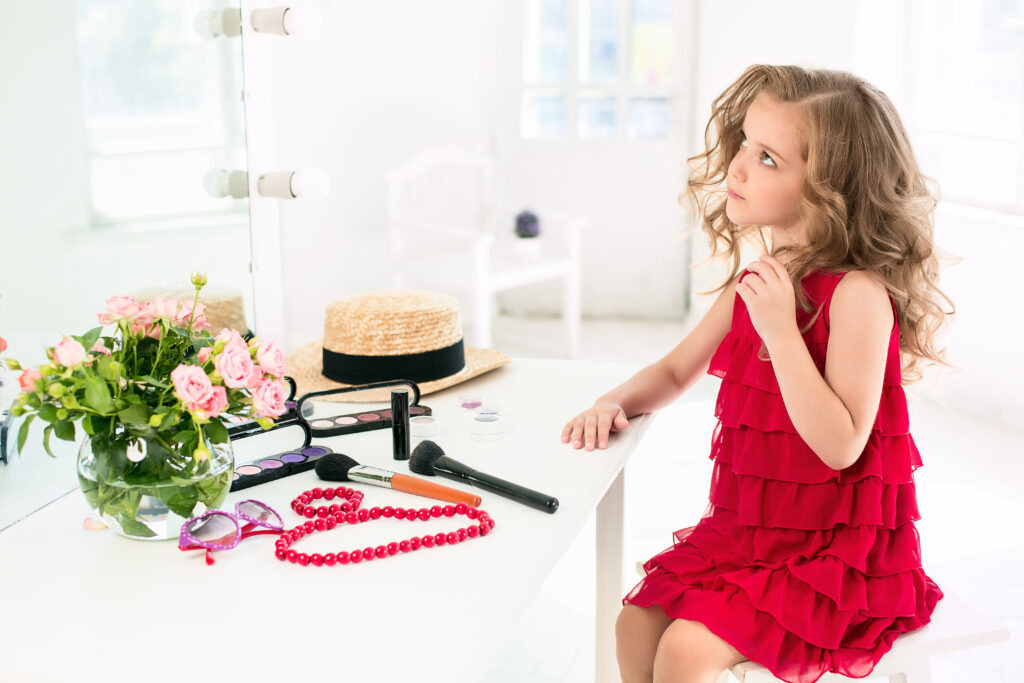 A little girl with red dress and cosmetics. She is in mother's bedroom, sitting near the mirror.